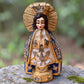 A Milagros figurine of the Virgin Mary Salud with a gold halo and crown, blue and beige robes, and silver-toned decorative details, standing on a stone surface with a blurred green outdoor background.