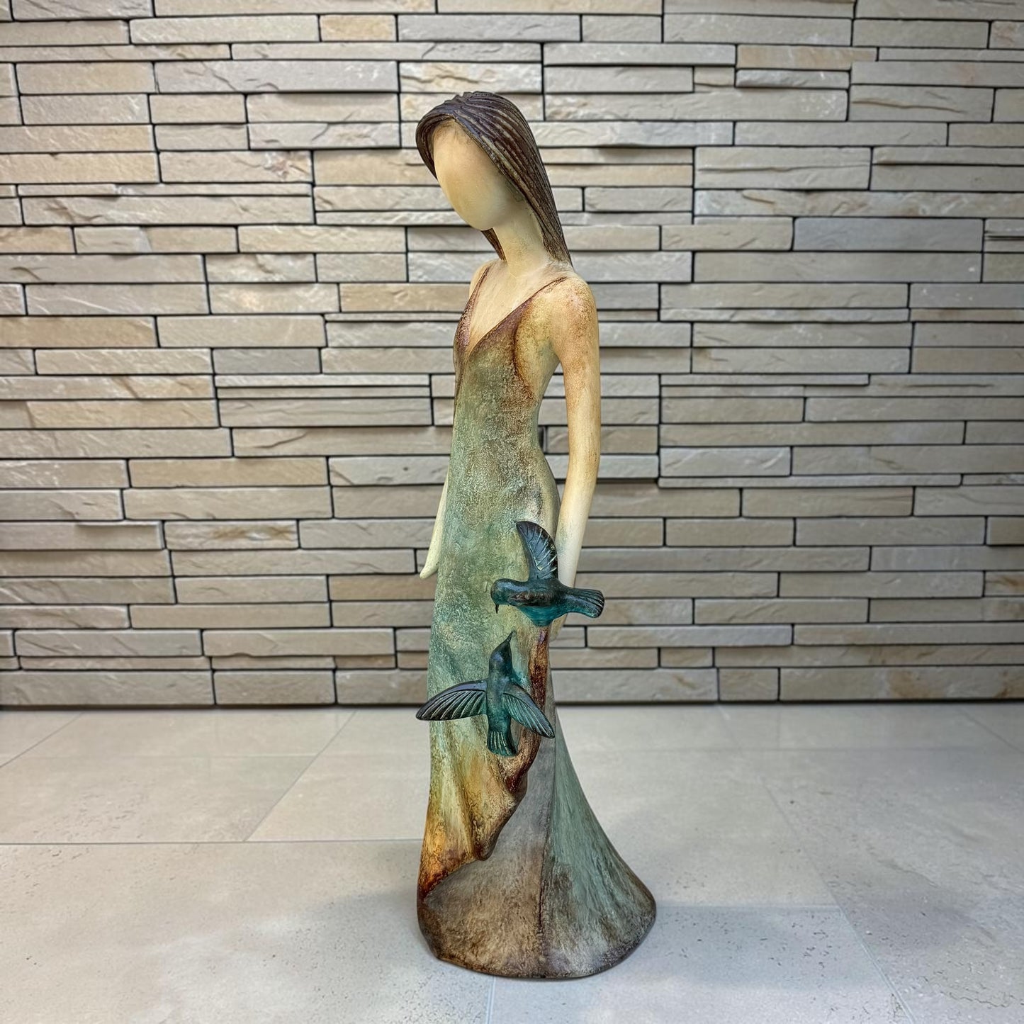 A Loretta Bertoli statue of a faceless woman in a long flowing dress with two hummingbirds, displayed against a stacked stone tile wall on a light tiled floor. This is a right side view. 
