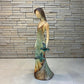 A Loretta Bertoli statue of a faceless woman in a long flowing dress with two hummingbirds, displayed against a stacked stone tile wall on a light tiled floor. This is a right side view. 
