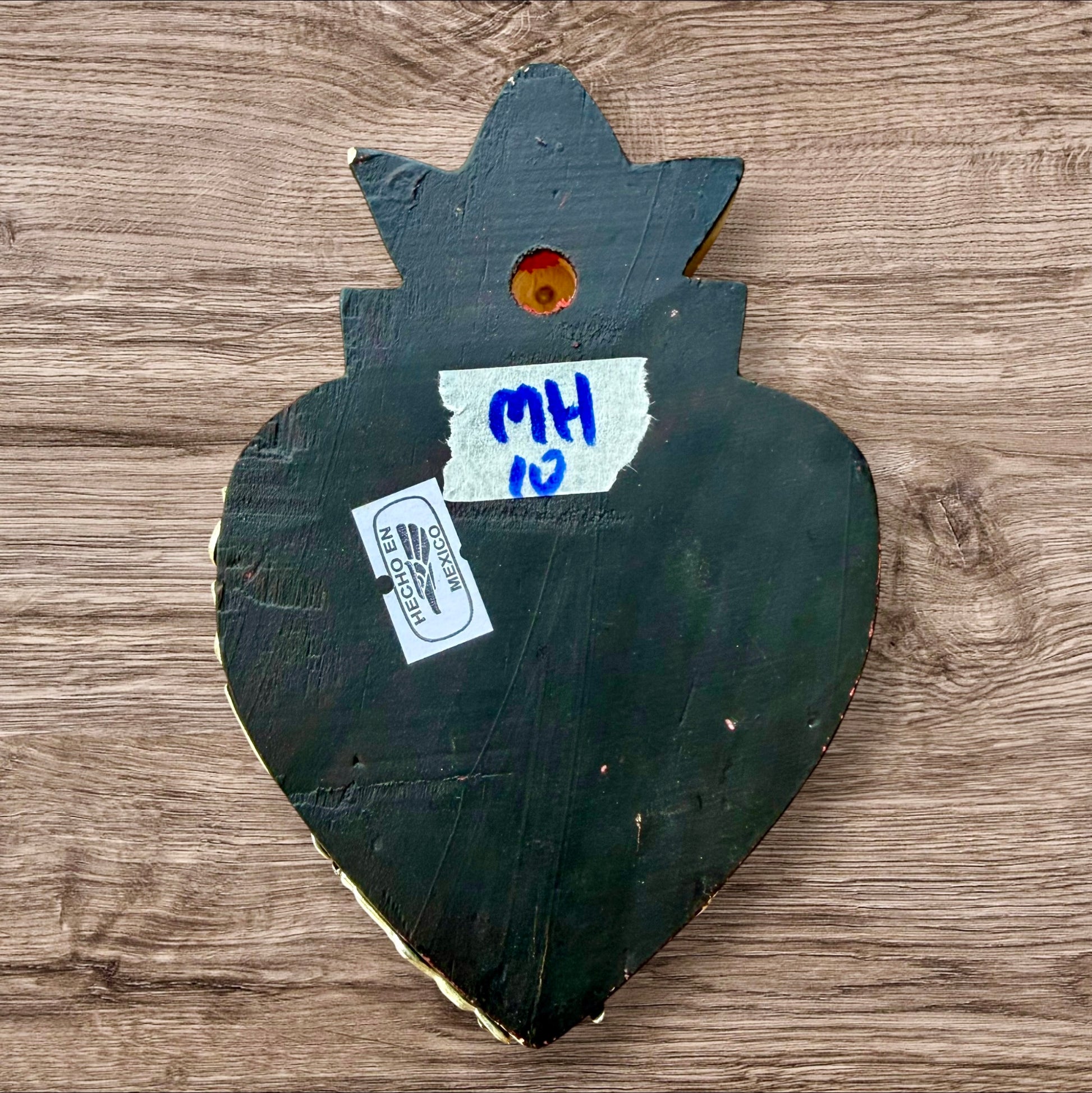 A Milagros heart featuring a gold textured top and a black base covered in raised silver-toned charms displayed on a wooden wall, back view.