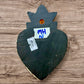 A Milagros heart featuring a gold textured top and a black base covered in raised silver-toned charms displayed on a wooden wall, back view.