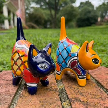 Two colorful talavera ceramic cat figurines standing on a brick surface outdoors with grass and trees blurred in the background. One cat is dark blue with orange, yellow, and red patterns, and the other is orange with blue, red, yellow, and white patterns.