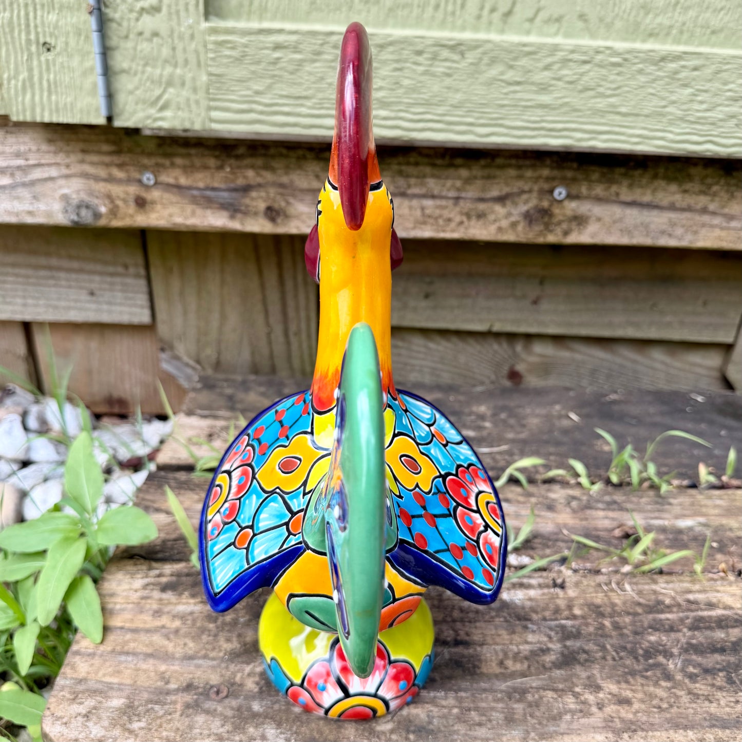 A colorful hand-painted ceramic Mexican-style rooster figurine standing on a rustic wooden surface outdoors, back tail view.