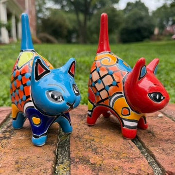 Two hand-painted Mexican-style ceramic cat figurines, one blue and one red, decorated with colorful geometric patterns, standing on a brick surface outdoors.