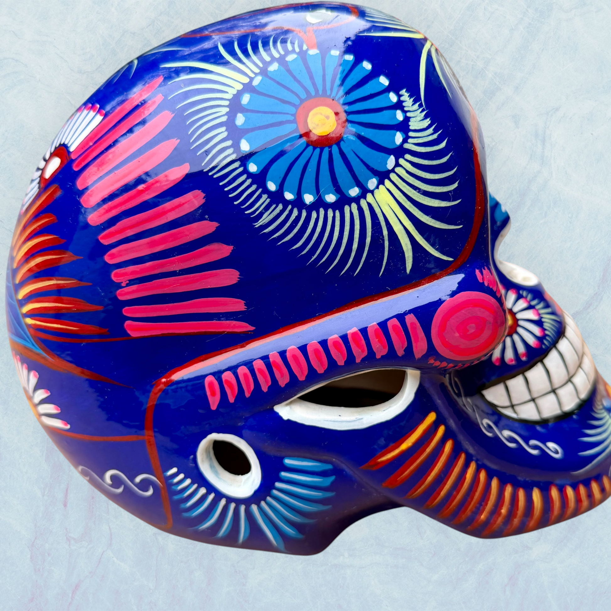 A glossy blue Day of the Dead sugar skull XGrande decorated with vibrant pink, blue, white, and orange floral and geometric patterns, shown against a light blue textured background, left side view.