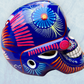 A glossy blue Day of the Dead sugar skull XGrande decorated with vibrant pink, blue, white, and orange floral and geometric patterns, shown against a light blue textured background, left side view.