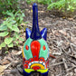 A colorful glazed ceramic cat figurine standing on mulch outdoors, with green plants blurred in the background. The cat is decorated with blue, orange, yellow, and white patterns, back of cat view.