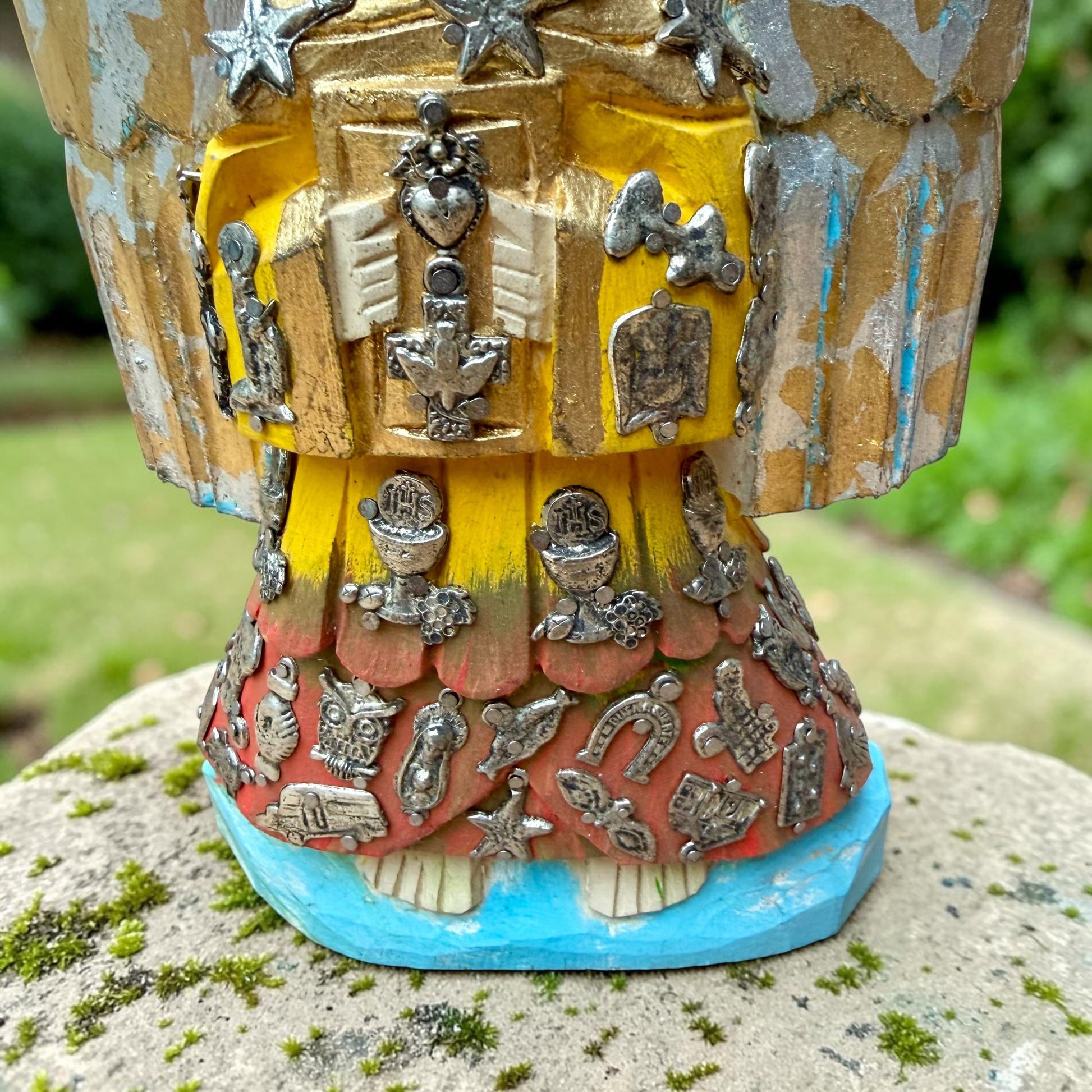 A handcrafted wooden Milagros angel figurine with large wings, decorated with metallic silver charms and painted in yellow, and orange, standing on a mossy stone with a blurred green garden background. Close up of lower robe and feet.