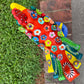 A colorful decorative rooster figurine standing on a brick surface outdoors, with a grassy lawn and trees in the background. The rooster is primarily red and yellow with many small multicolored flowers and painted details across its body and tail. Top view showing hand crafted flowers on the rooster.