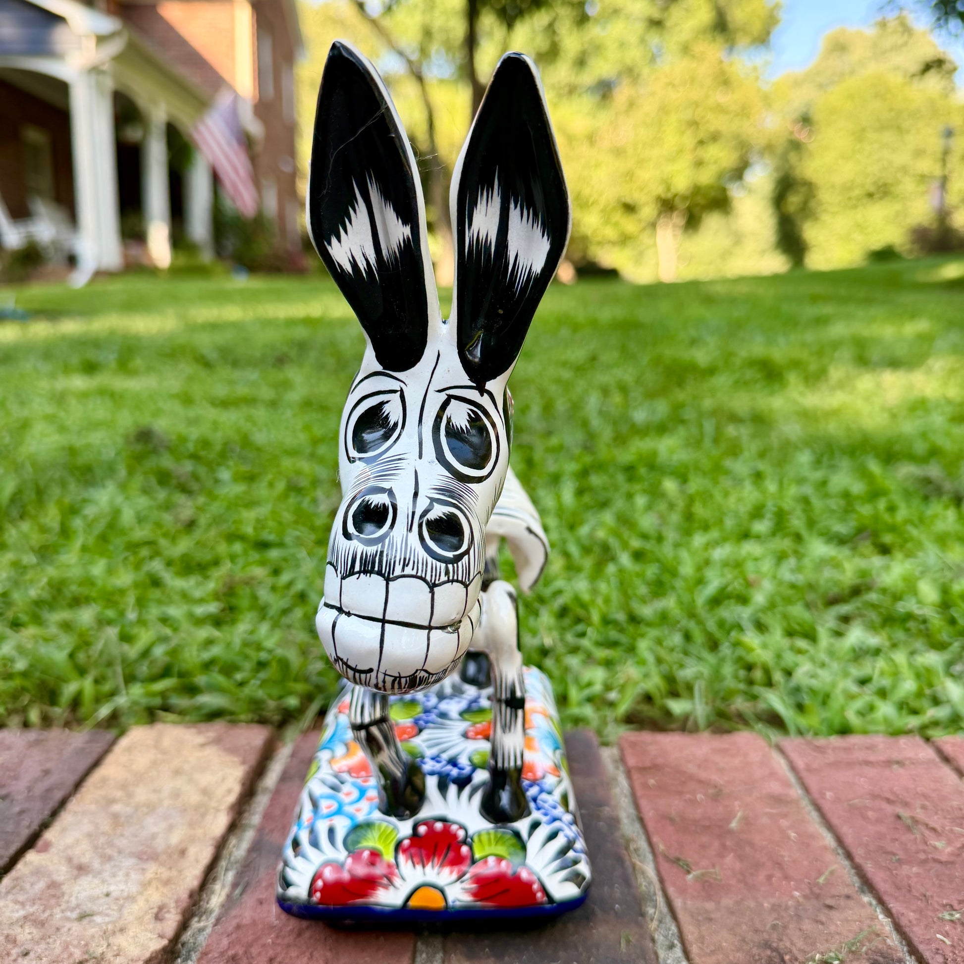 A colorful ceramic donkey Catrina figurine with a skeletal white-and-black design and a floral base, photographed outdoors on a brick surface with a blurred green lawn and house in the background. This is the front face view.