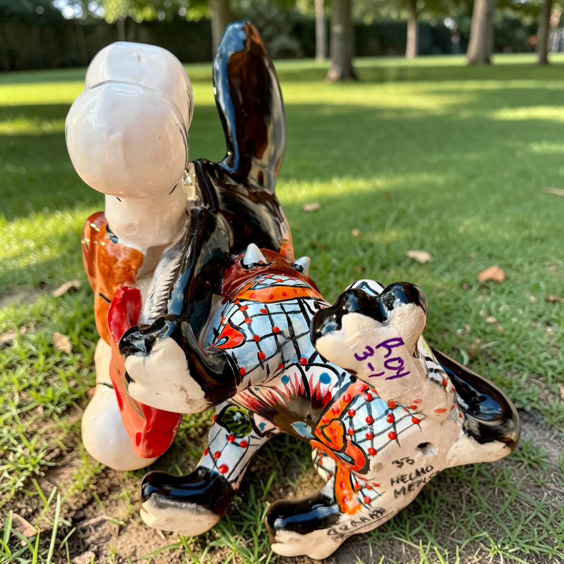 A colorful Talavera dog statue holding a big white bone, photographed outdoors on grass with trees in the background. The dog has a glossy black body, blue eyes, an orange snout, and multicolored patterned ears.  Bottom view showing artist signature.