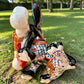 A colorful Talavera dog statue holding a big white bone, photographed outdoors on grass with trees in the background. The dog has a glossy black body, blue eyes, an orange snout, and multicolored patterned ears.  Bottom view showing artist signature.