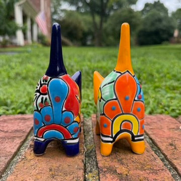 Two colorful talavera ceramic cat figurines standing on a brick surface outdoors with grass and trees blurred in the background. One cat is dark blue with orange, yellow, and red patterns, and the other is orange with blue, red, yellow, and white patterns. back view showing pointy tail