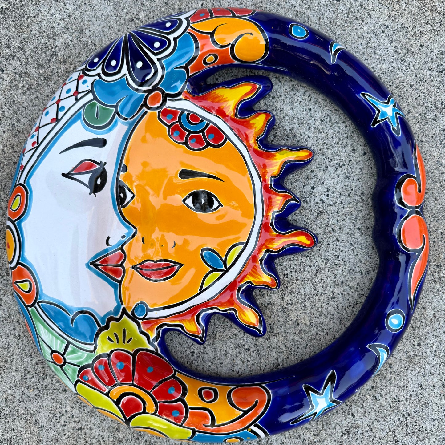 A colorful hand-painted ceramic wall plaque featuring a crescent moon and sun design, set against a gray stone background.