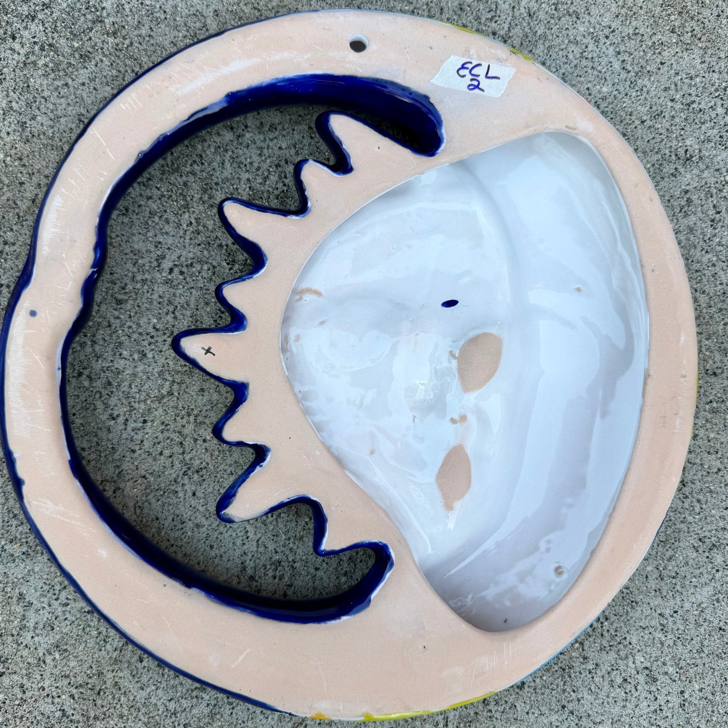 A colorful hand-painted talavera wall hanging shaped like a crescent moon and sun, featuring a glossy finish and vibrant orange, blue, yellow, and white details, displayed on a gray textured concrete surface. This is a photo of the back view featuring the hanging hole.