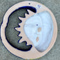 A colorful hand-painted talavera wall hanging shaped like a crescent moon and sun, featuring a glossy finish and vibrant orange, blue, yellow, and white details, displayed on a gray textured concrete surface. This is a photo of the back view featuring the hanging hole.
