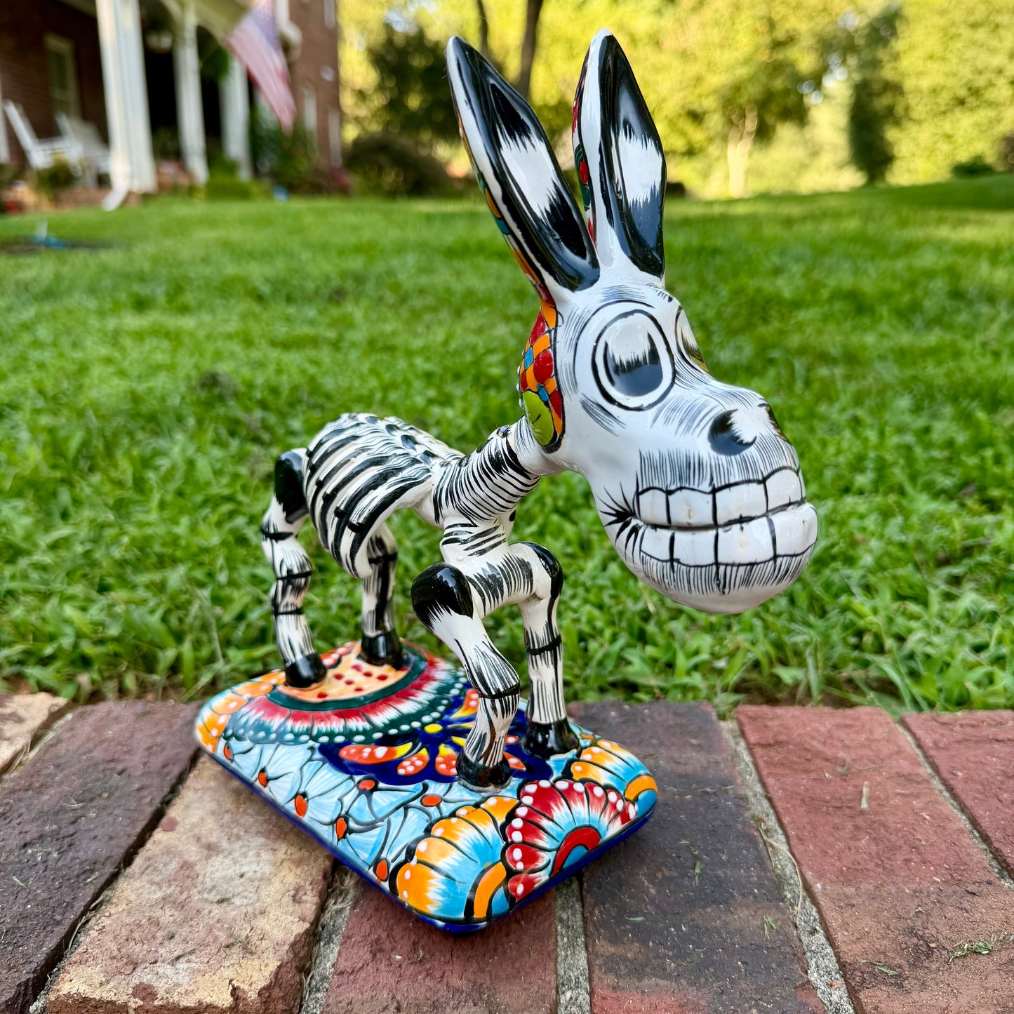 A colorful Catrina donkey figurine standing on a decorated base, photographed outdoors on a brick surface with a grassy yard and house in the background.