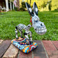 A colorful Catrina donkey figurine standing on a decorated base, photographed outdoors on a brick surface with a grassy yard and house in the background.