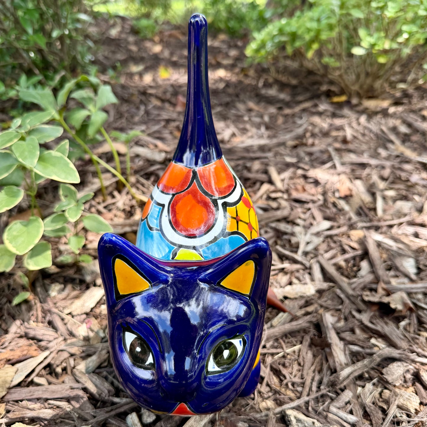 A colorful glazed ceramic cat figurine standing on mulch outdoors, with green plants blurred in the background. The cat is decorated with blue, orange, yellow, and white patterns, front face view.