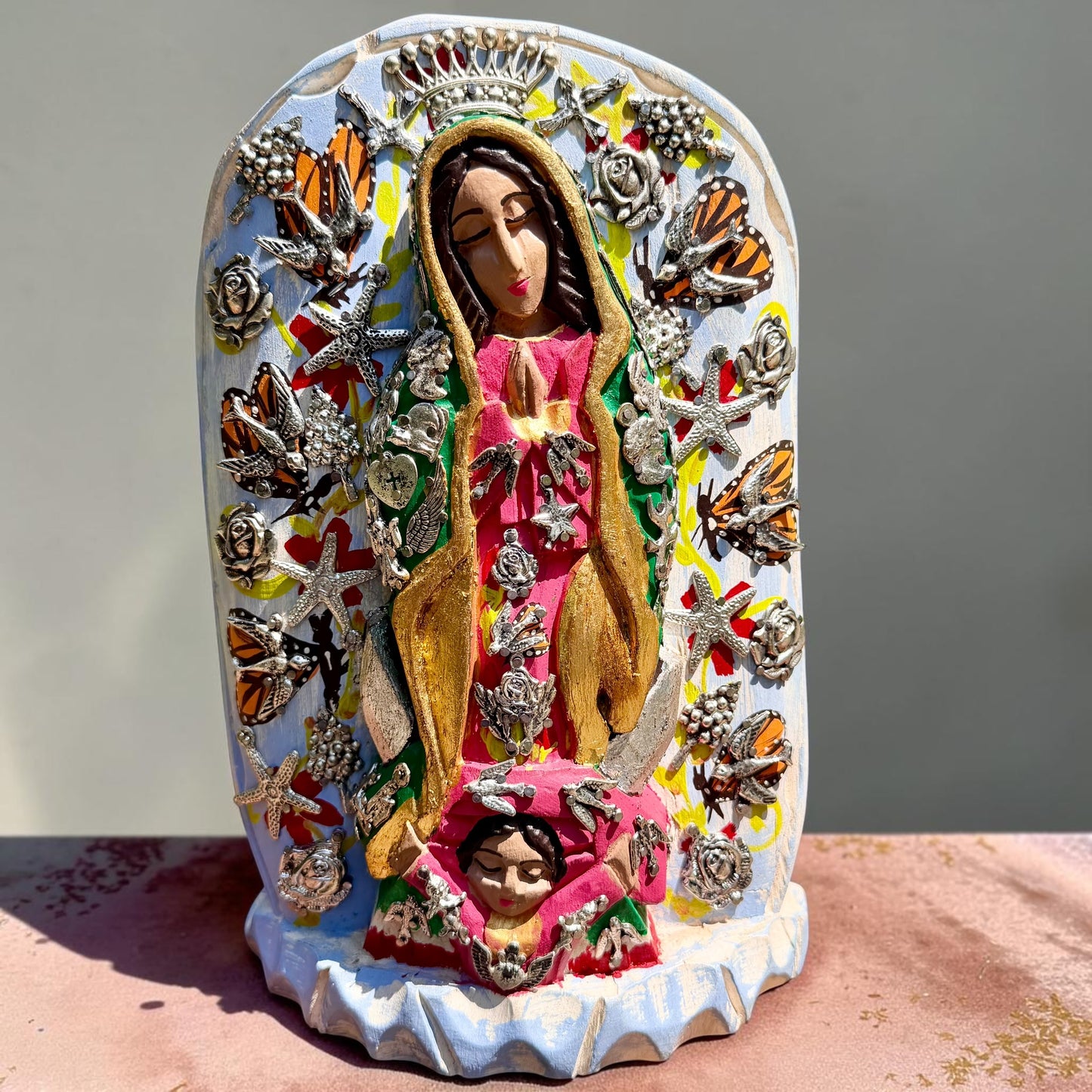 A colorful religious milagros wood figurine of Our Lady of Guadalupe surrounded by butterflies, displayed on a pink surface against a light blue background. 