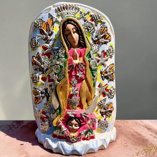 A colorful religious milagros wood figurine of Our Lady of Guadalupe surrounded by butterflies, displayed on a pink surface against a light blue background. 