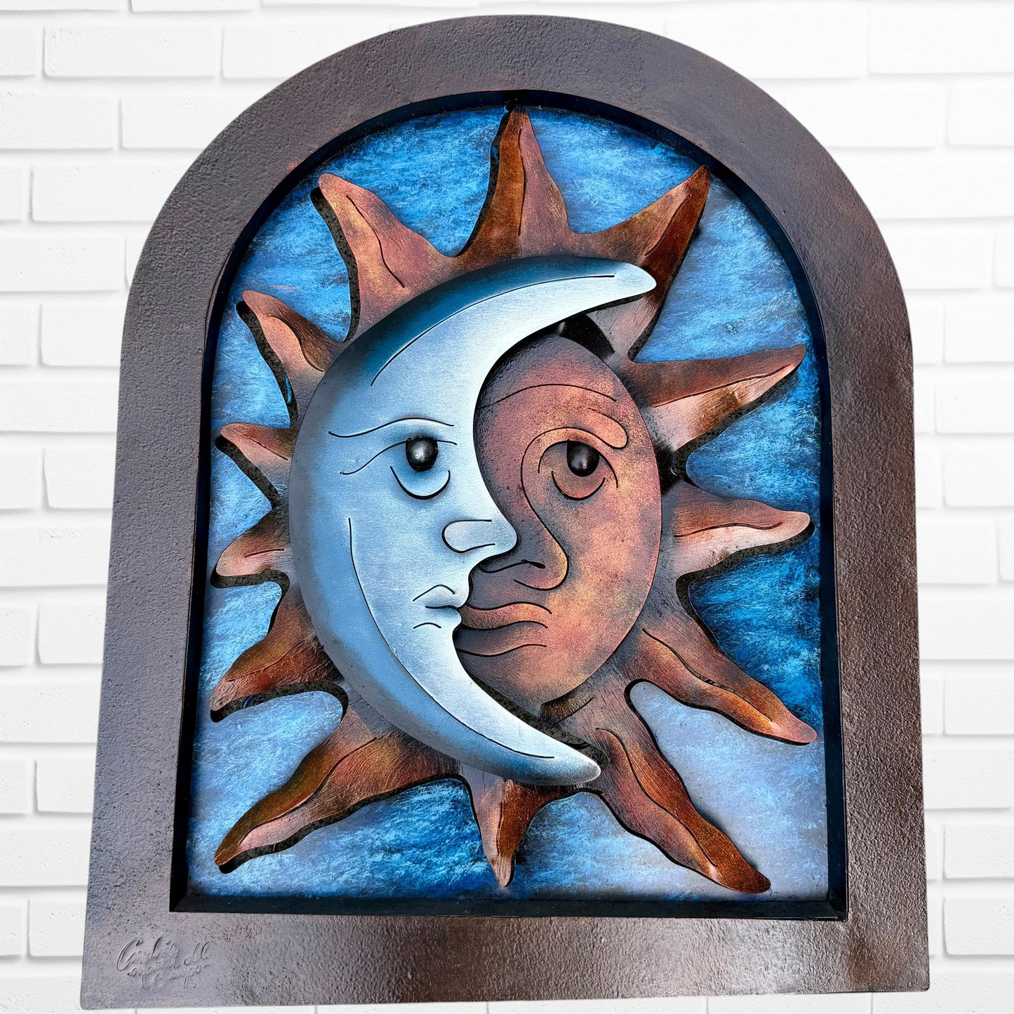 Decorative wall art of a sun and moon face with a metal frame on a white brick wall. Designed by artist Andres Martin.