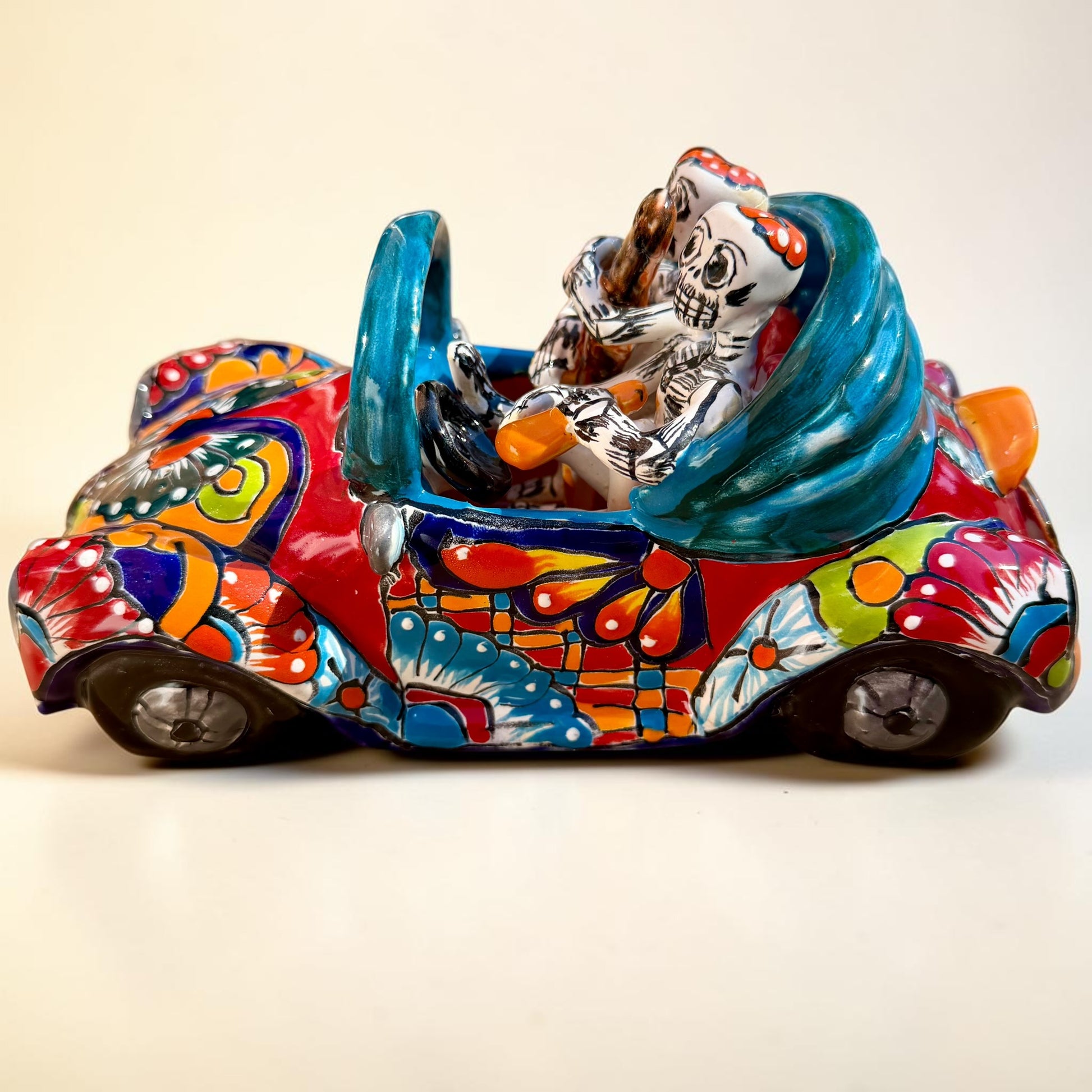A colorful hand-painted ceramic Mexican-style convertible car figurine with Day of the Dead skeleton characters inside, shown on a neutral beige background. Right side view of the car.