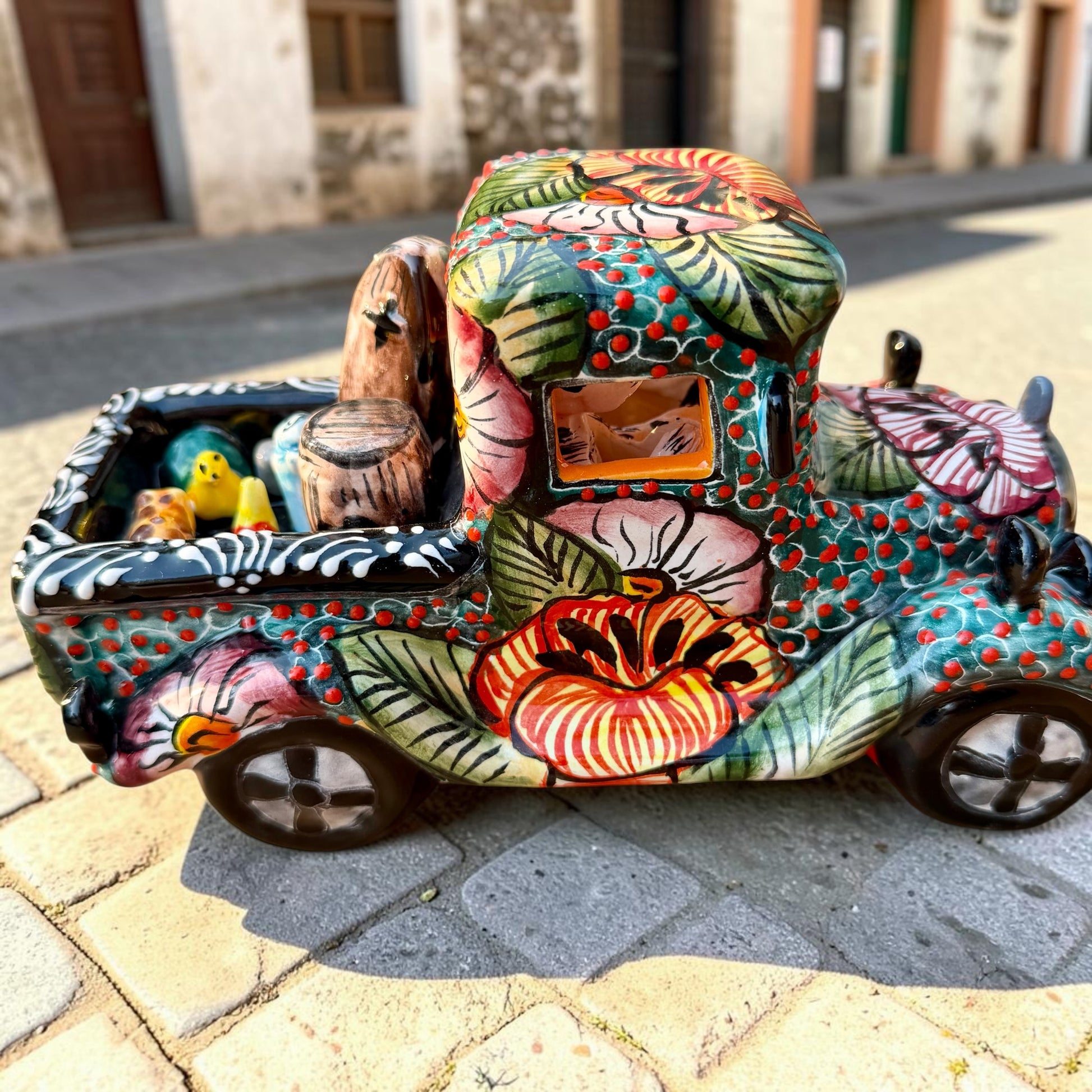 A decorative Talavera day of the dead pick up truck figurine two skeleton passengers. The truck is vibrant, with a hand-painted tropical floral pattern, photographed outdoors on a stone-paved street. This is the left side view of the pick up truck.