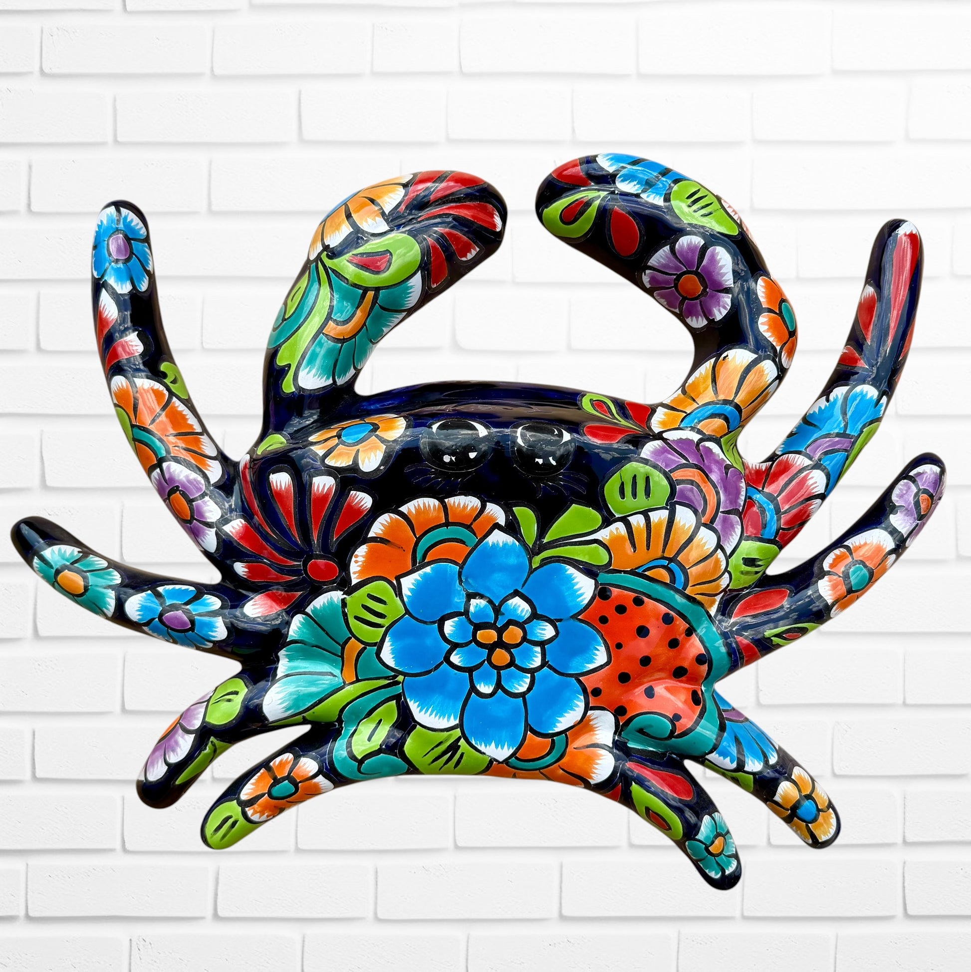 A colorful talavera crab wall art sculpture decorated with vibrant floral patterns, shown against a white brick wall background, top view of crab.