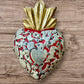 A Milagros heart featuring a gold textured top and a red base covered in raised silver-toned charms displayed on a wooden wall.