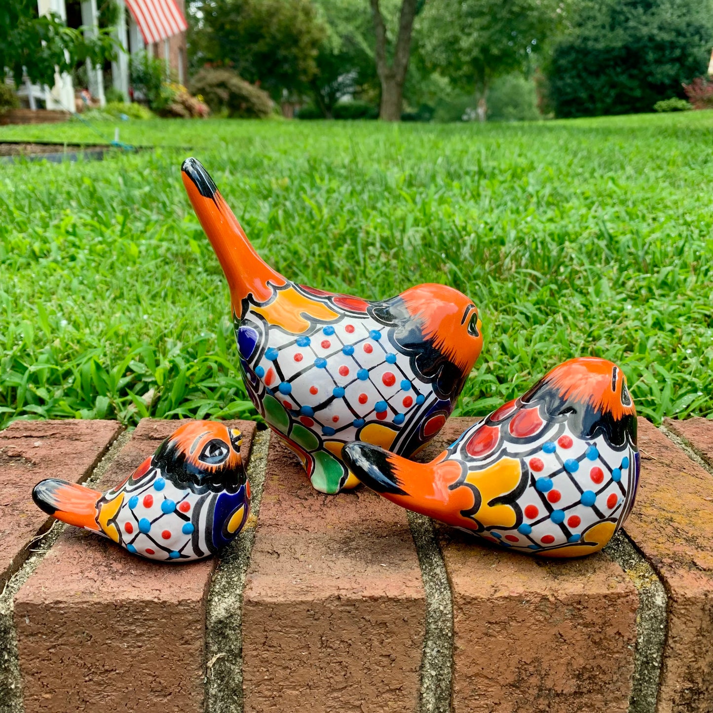 Three hand-painted ceramic bird garden statues in orange, blue, yellow, and black, displayed on a brick ledge in a backyard with a green lawn and trees in the background left side view