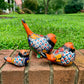 Three hand-painted ceramic bird garden statues in orange, blue, yellow, and black, displayed on a brick ledge in a backyard with a green lawn and trees in the background left side view