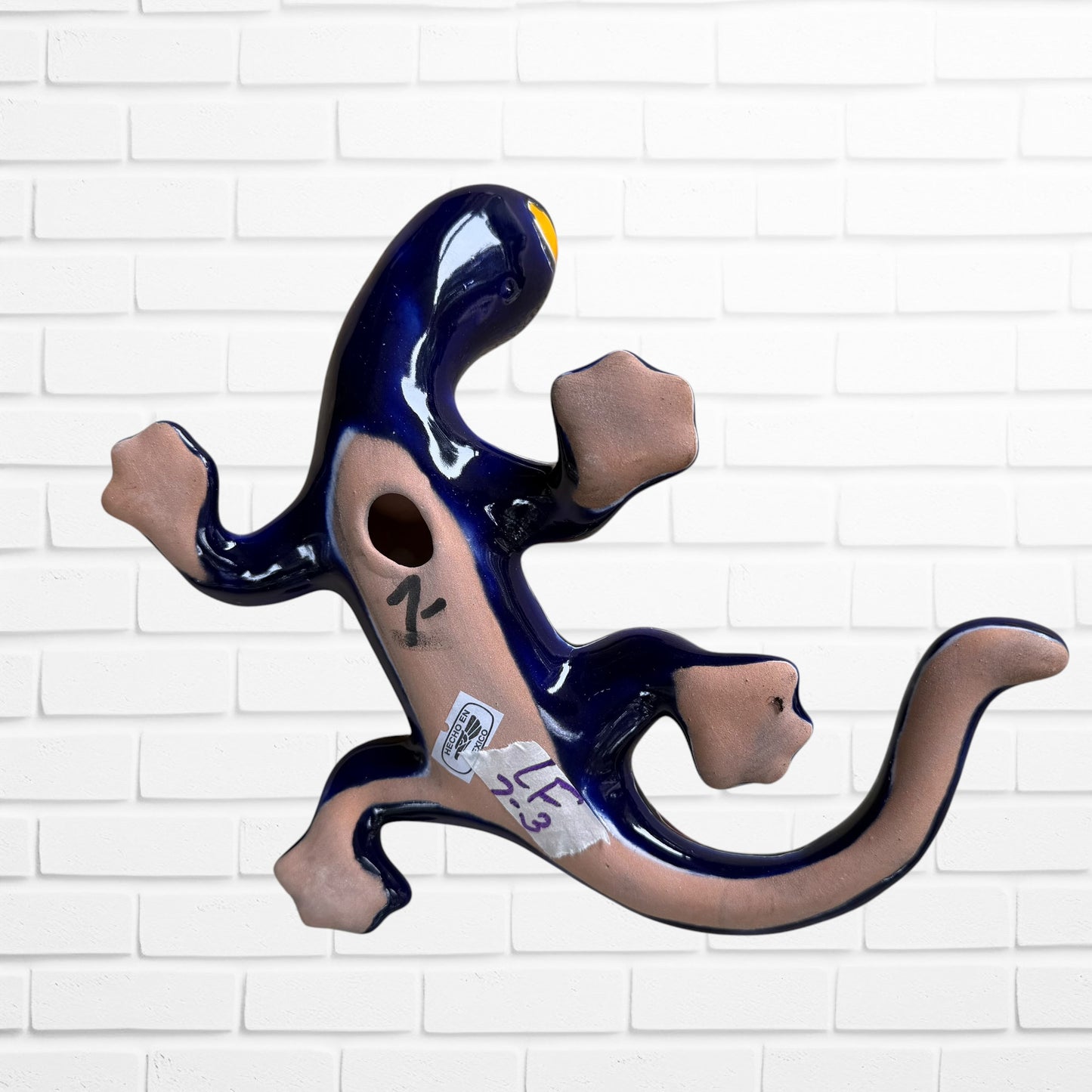 A colorful glazed ceramic lizard wall sculpture with a vibrant multicolor pattern, displayed against a white brick wall background bottom featuring hanging hole.