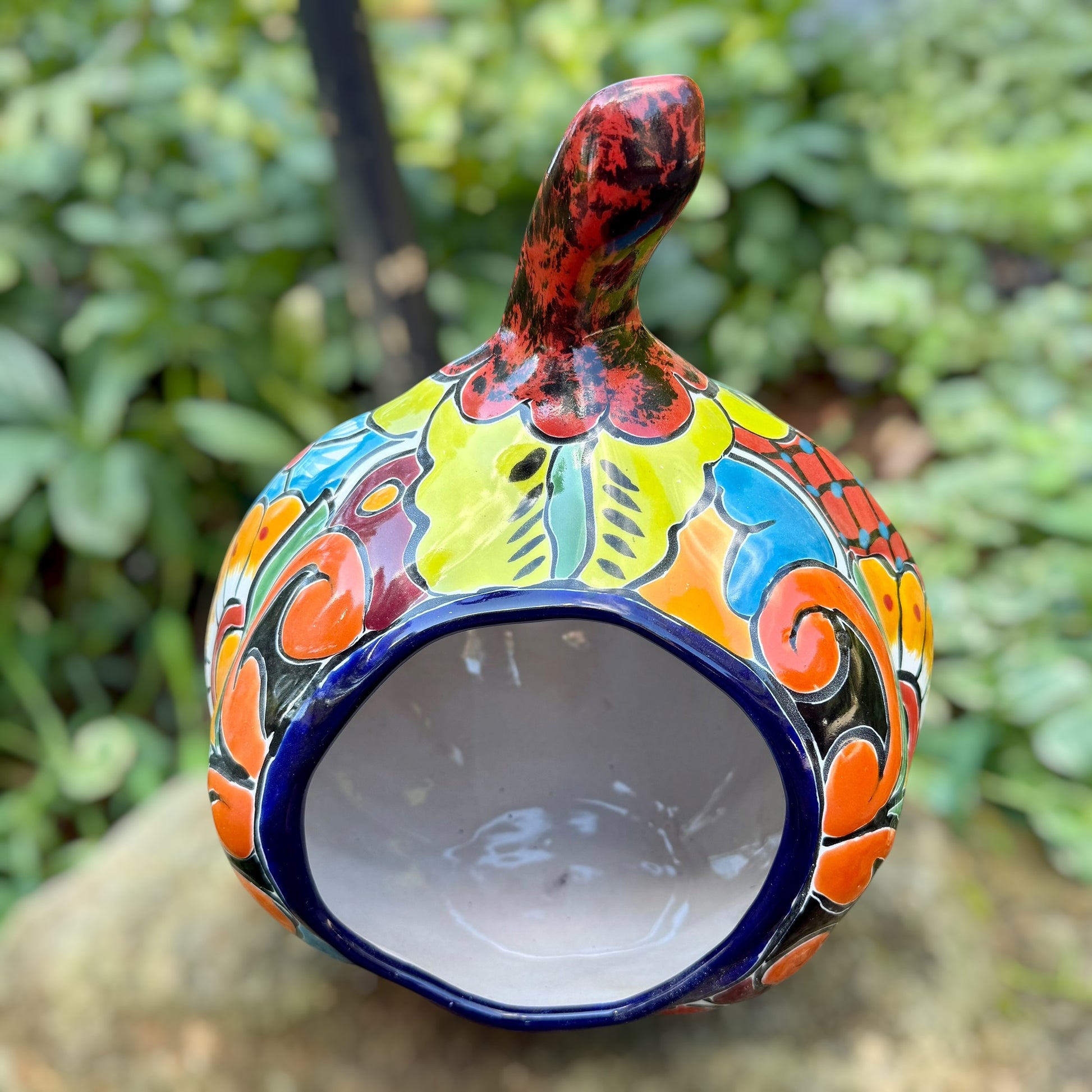 A hand-painted ceramic jack-o'-lantern pumpkin with a red face, black carved eyes and mouth, and a multicolored floral pattern on a garden background. Back of pumpkin with opening.