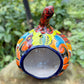 A hand-painted ceramic jack-o'-lantern pumpkin with a red face, black carved eyes and mouth, and a multicolored floral pattern on a garden background. Back of pumpkin with opening.