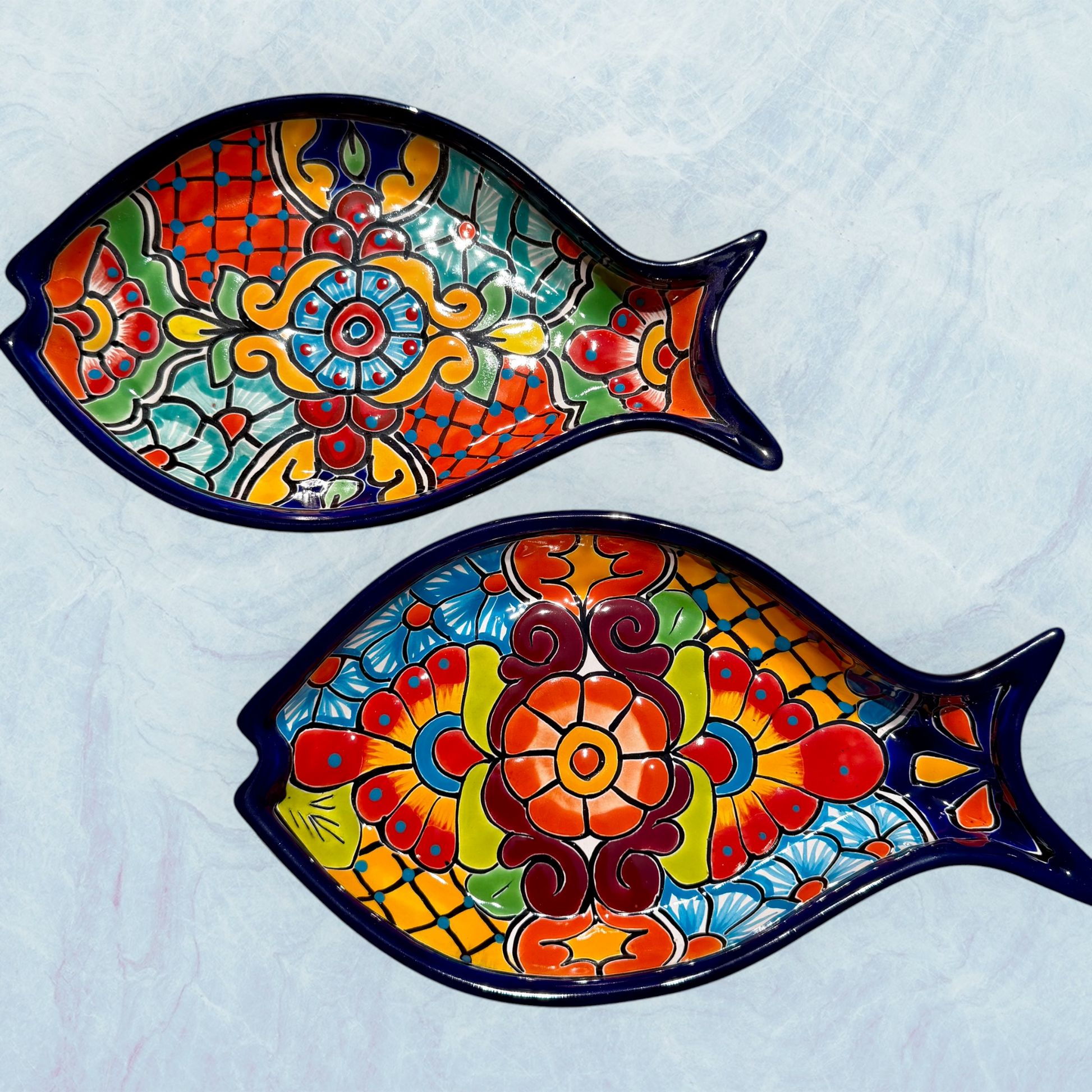 Two decorative blue fish-shaped ceramic plates with vibrant hand-painted floral and geometric patterns in red, orange, yellow, green, blue, and purple, set against a light blue background.