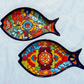 Two decorative blue fish-shaped ceramic plates with vibrant hand-painted floral and geometric patterns in red, orange, yellow, green, blue, and purple, set against a light blue background.
