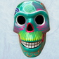 A decorative skull sculpture with intricate colorful sugar skull style patterns painted on a teal colored base, set against a light blue textured background, front view.