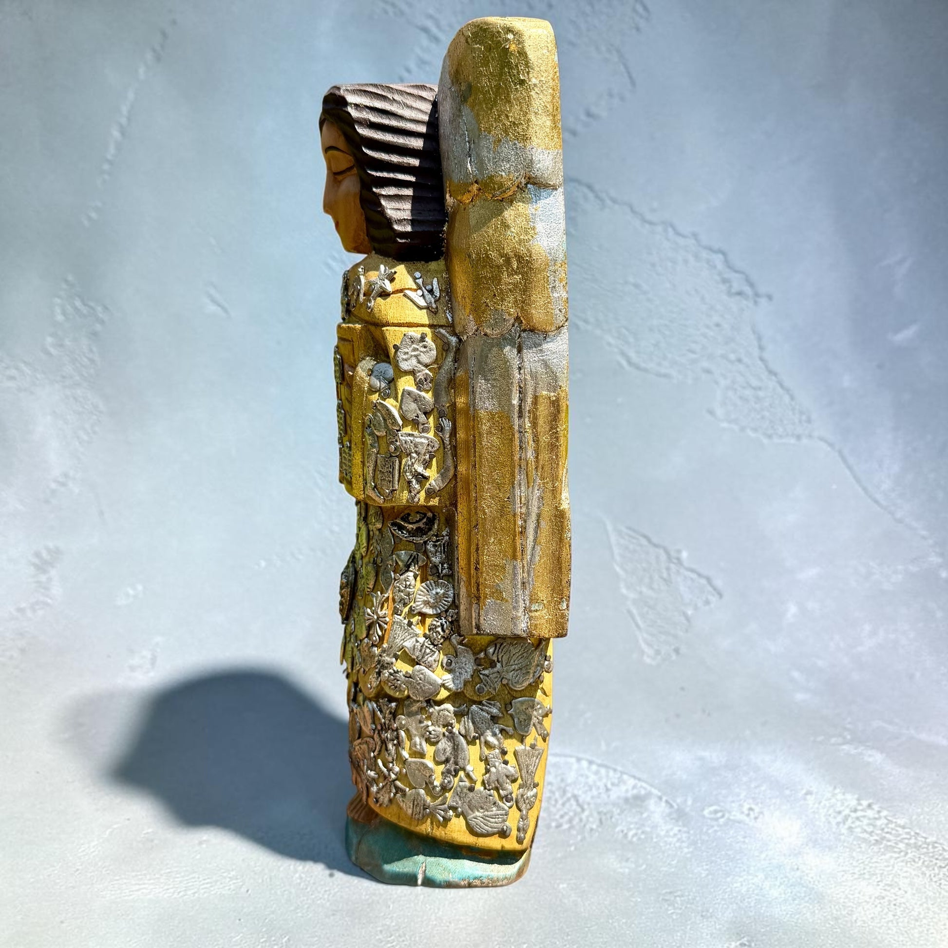 Milagros wooden guardian angel figurine wearing a yellow robe with gold and silver metallic details, standing on a light blue textured background, right side view.