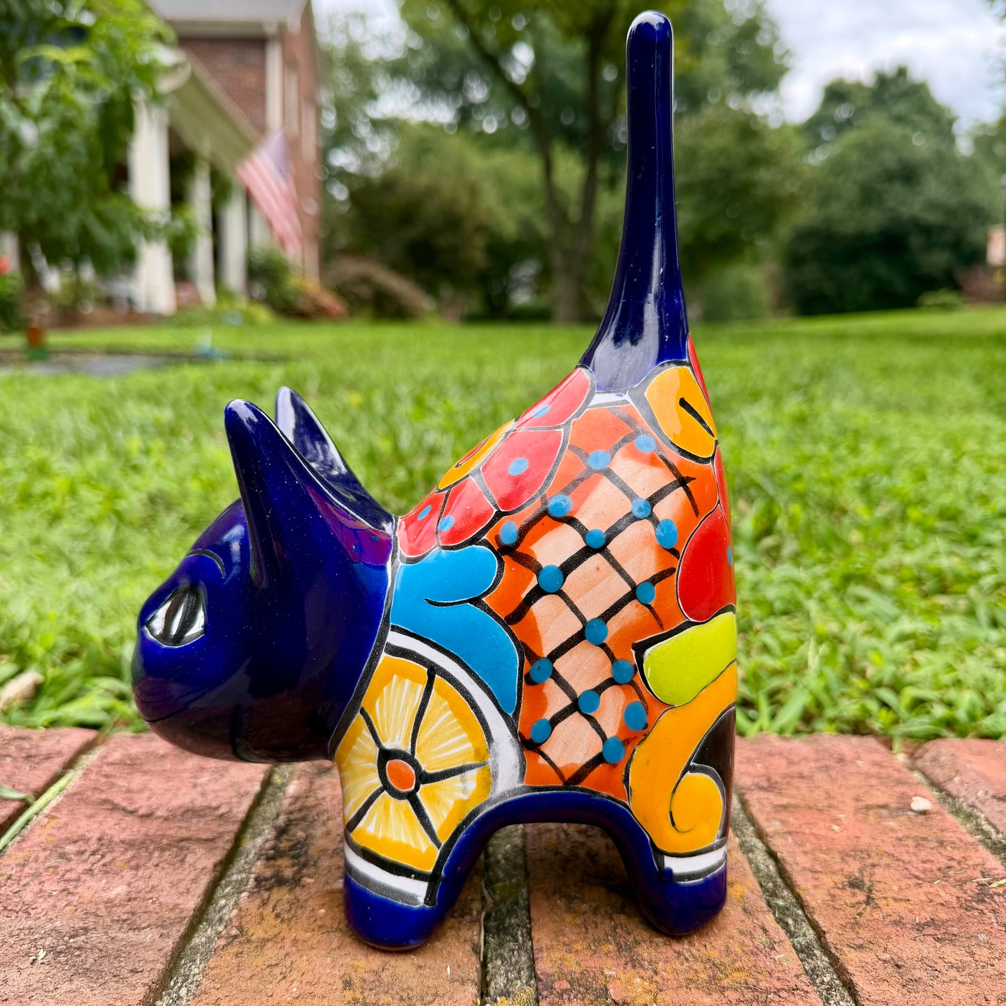 A glossy talavera ceramic cat figurine decorated with vibrant hand-painted patterns in blue, orange, yellow, and red, standing on a brick path with a green lawn and a house in the blurred background. Right side view.