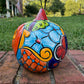 A hand-painted Talavera  jack-o'-lantern pumpkin sits on a brick patio with a green grass lawn and trees in the background. The pumpkin features a glossy orange base with hand-painted details in red, yellow, blue, and green. Right side of the pumpkin.