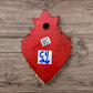 A Mexican Milagros heart in red with a gold flaming top, covered in silver charms, back view. 