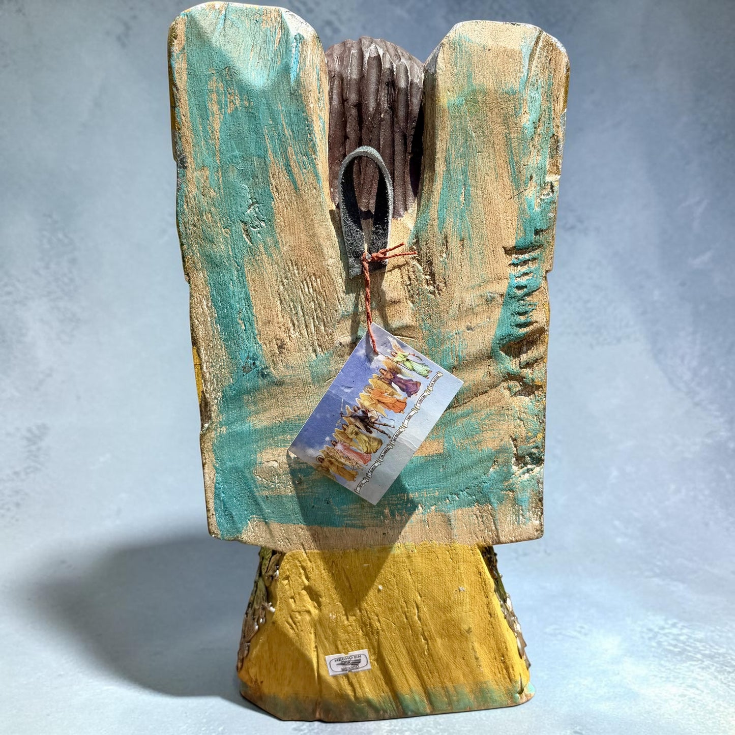 Milagros wooden guardian angel figurine wearing a yellow robe with gold and silver metallic details, standing on a light blue textured background, back view of wings Showing loop for hanging.