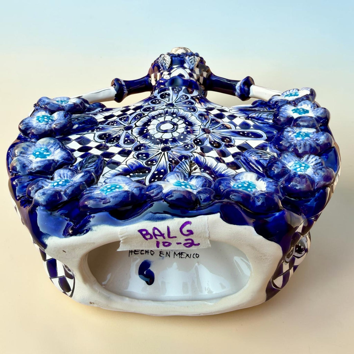 A Day of the Dead style talavera figurine of a skeleton woman in a wide floral skirt, painted in deep blue, black, white, and light blue on a soft beige-to-blue gradient background. Bottom showing artist signature.
