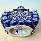 A Day of the Dead style talavera figurine of a skeleton woman in a wide floral skirt, painted in deep blue, black, white, and light blue on a soft beige-to-blue gradient background. Bottom showing artist signature.