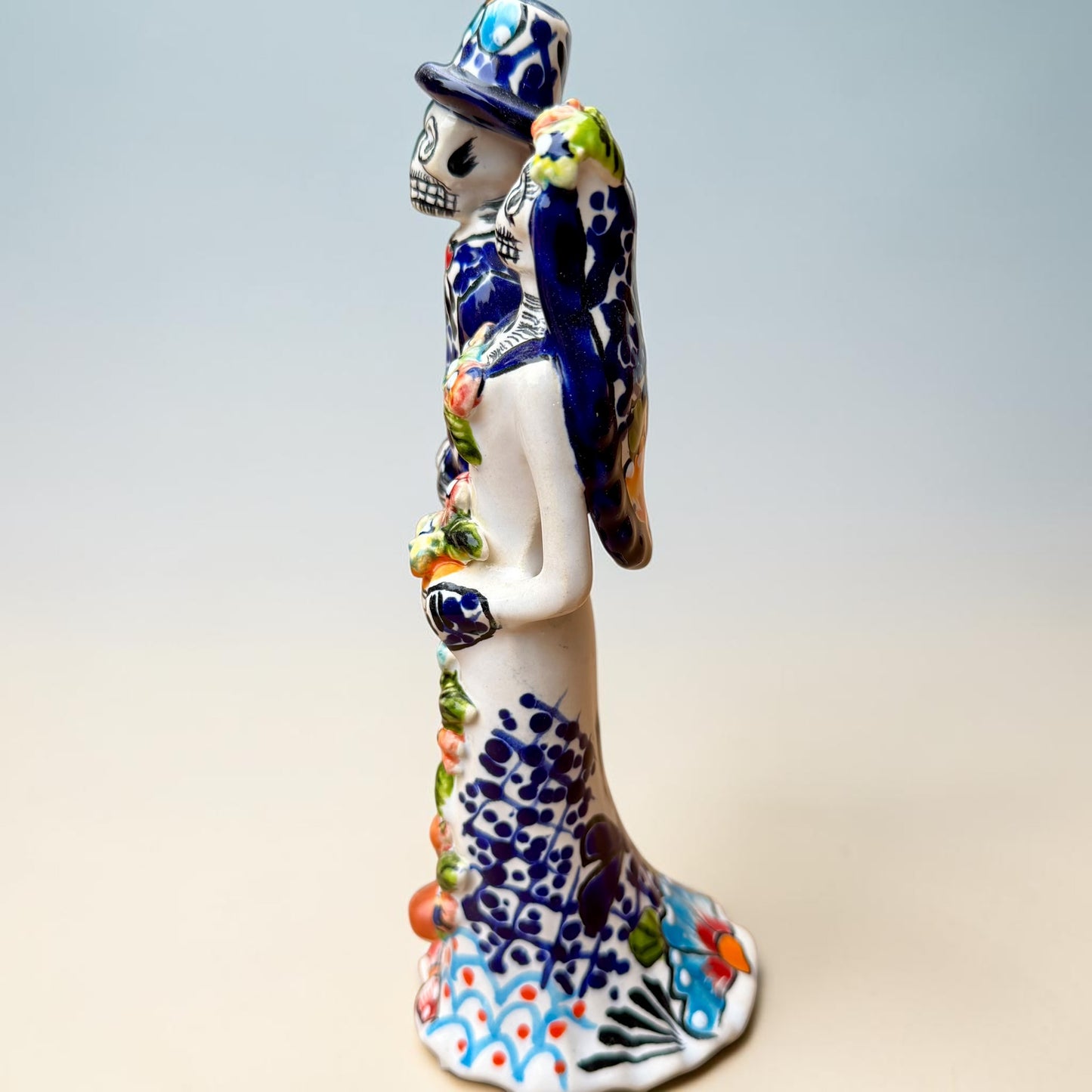 A hand-painted Talavera Day of the Dead bride and groom figurine. The groom is dressed in a blue suit with white patterns, a black bow tie, and an orange-red bow tie accent, wearing a blue hat. Right side view of couple featuring bride.