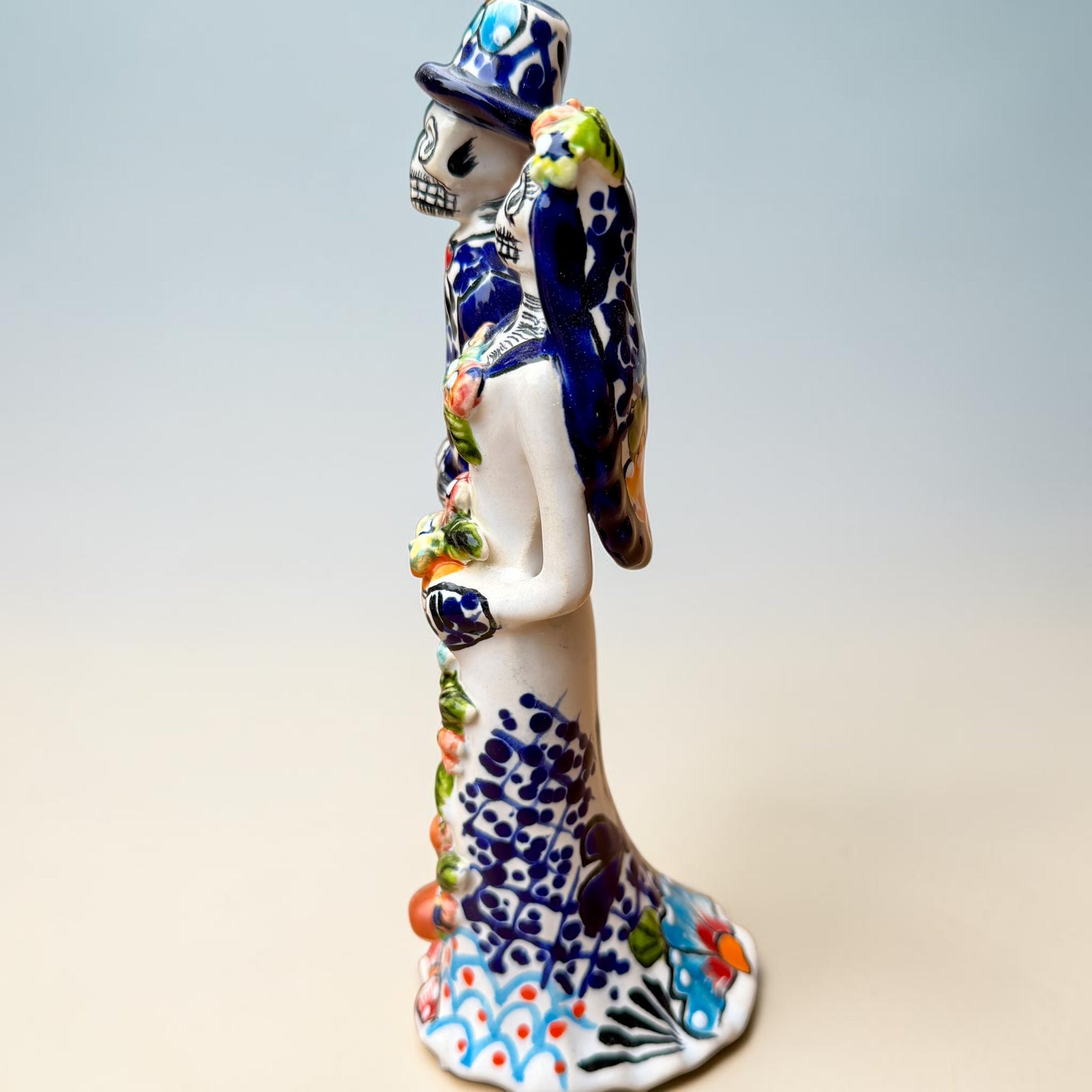 A hand-painted Talavera Day of the Dead bride and groom figurine. The groom is dressed in a blue suit with white patterns, a black bow tie, and an orange-red bow tie accent, wearing a blue hat. Right side view of couple featuring bride.