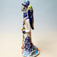 A hand-painted Talavera Day of the Dead bride and groom figurine. The groom is dressed in a blue suit with white patterns, a black bow tie, and an orange-red bow tie accent, wearing a blue hat. Right side view of couple featuring bride.