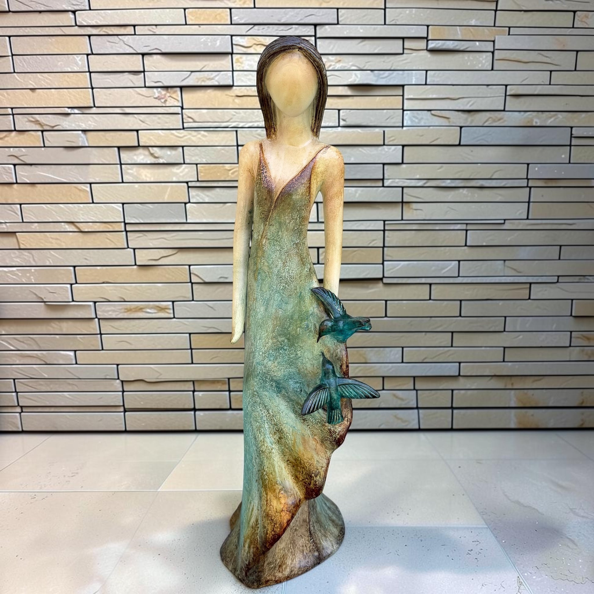 A Loretta Bertoli statue of a faceless woman in a long flowing dress with two hummingbirds, displayed against a stacked stone tile wall on a light tiled floor.