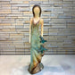 A Loretta Bertoli statue of a faceless woman in a long flowing dress with two hummingbirds, displayed against a stacked stone tile wall on a light tiled floor.